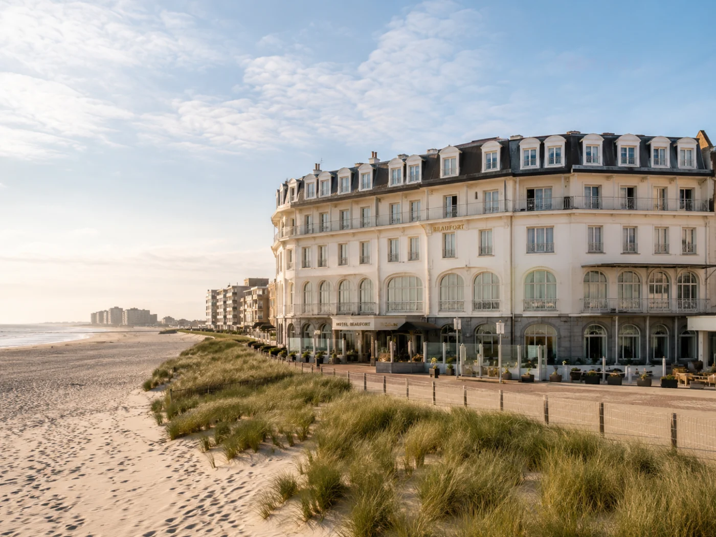 Statig wit belle-epoquehotel aan de Belgische kust met een breed zandstrand, duinen met helmgras en een wandelpromenade in zacht ochtendlicht