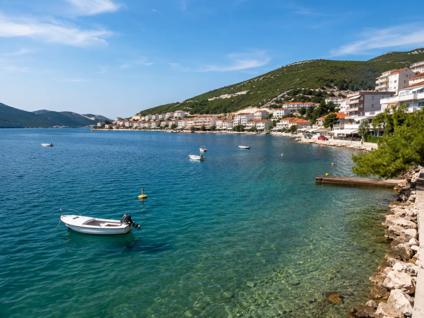 De kust van Neum in Bosnië en Herzegovina met helder turquoise water, kleine boten en een heuvel met witte huizen en oranje daken aan de Adriatische Zee