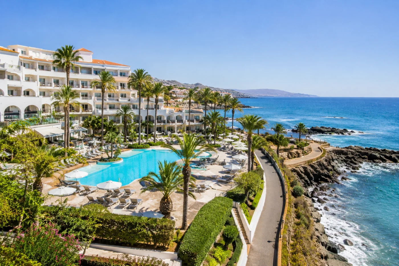 Wit resort aan de kust van Cyprus met een groot zwembad, palmen, ligbedden en een kustpad langs de rotsachtige zee