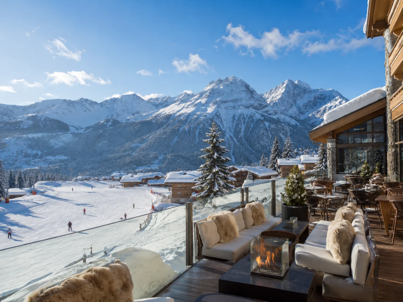 Sunny terrace of a winter sports hotel with lounge sofas and a fire pit right by the slopes, with snow-capped mountain peaks during the spring half-term